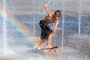 Junger Junge fährt mit dem Skateboard durch einen Springbrunnen mit Regenbogeneffekt