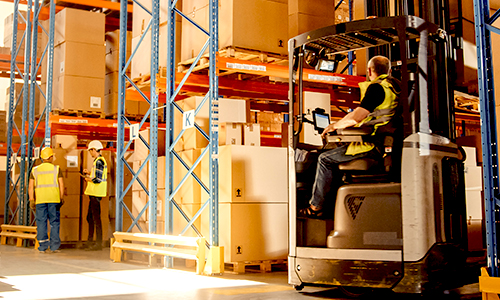 Fabriklager mit Regalen voller großer Kartons. Im Vordergrund benutzt ein Arbeiter ein Tablet auf einem Gabelstapler und zwei Arbeiter beraten sich im Hintergrund. 