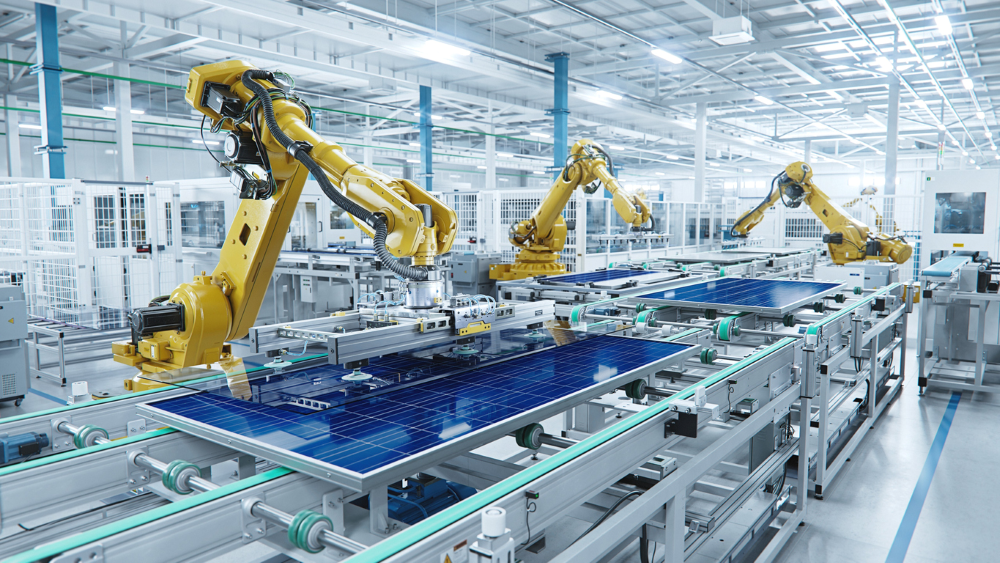 Several yellow robotic arms working on solar panels on a production line. The white overhead lights and white walls give the factory a very bright appearance.