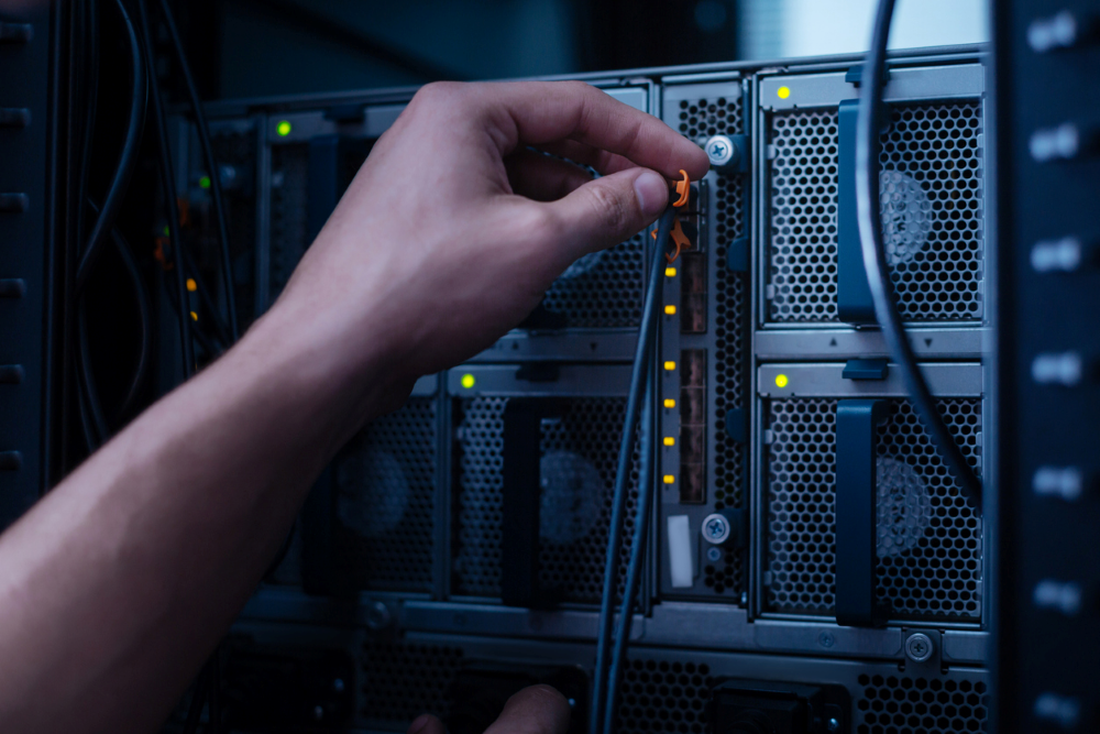 A hand inserts a small plugin into a computer port. Lights on the board indicate the device is on, demonstrating the use of hot swapping. The lighting is dark and cool-toned.