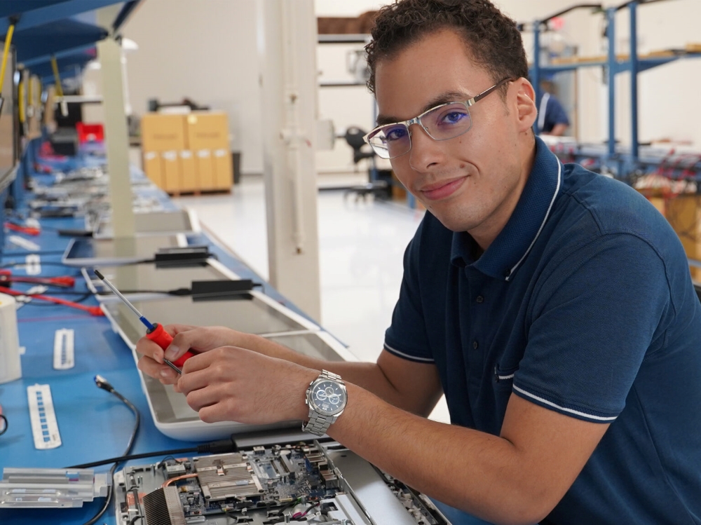 A smiling Teguar employee is working on a line of computers laid out on a work station.