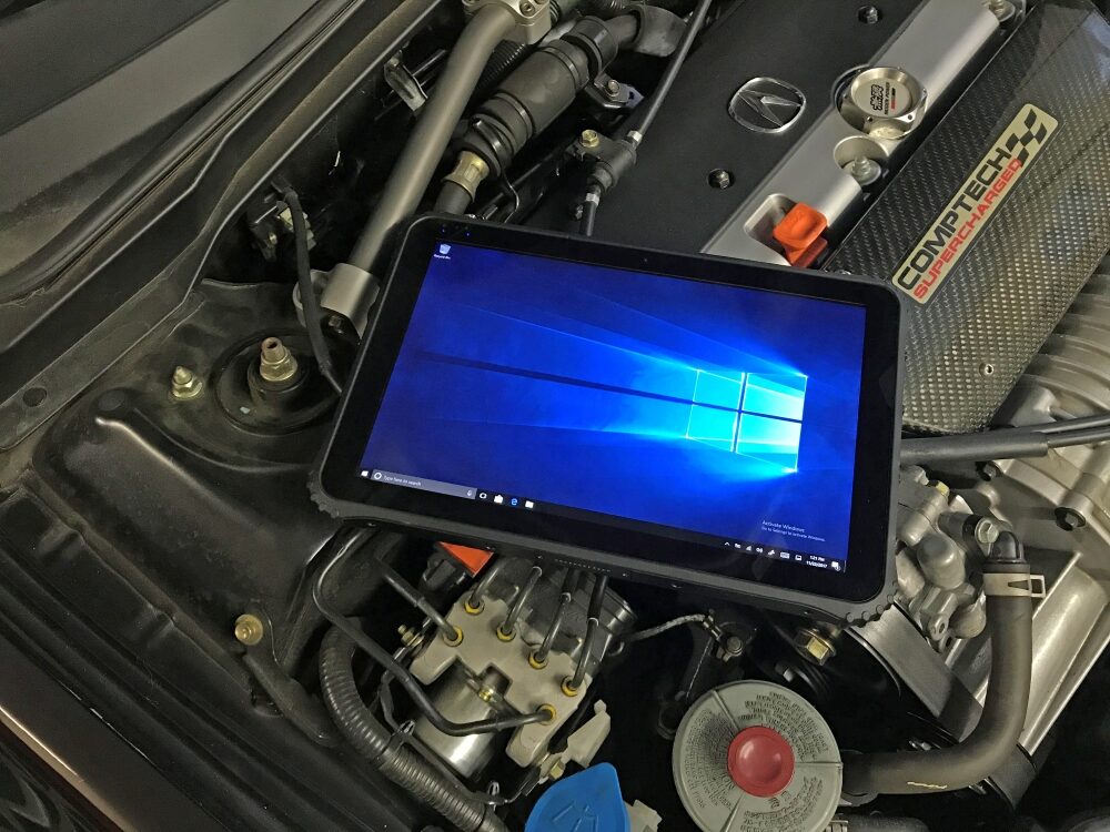 A rugged tablet opened to the Windows 10 home screen, resting on a car engine.