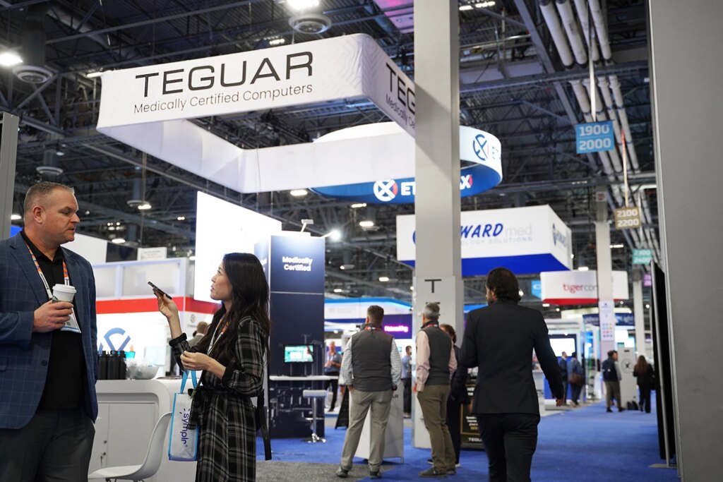 Großaufnahme des Teguar-Standes in der Ausstellungshalle. Einige Teilnehmer laufen herum und sprechen miteinander. 