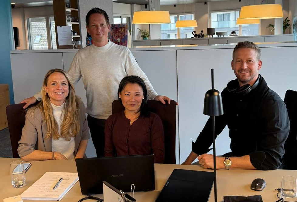 Vier Mitarbeiter von Teguar Europe posieren für ein Foto in den Büroräumen der Teguar AG. CEO Jonathan Staub steht in der hinteren Reihe, und Mitinhaberin Corinne Staub, Solutions Support Specialist Linda Deutinger und Teguar Europe Branch Manager Philipp Knupp sitzen vorne an einem Arbeitstisch. Alle lächeln und schauen in die Kamera.  