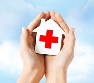 Cupped hands in front of a cloudy background holding a small white house with a red cross on it