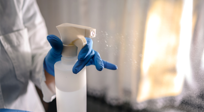 A gloved hand sprays a cleaning chemical from a spray bottle onto a surface out of view. The person is in a white coat and the room is softly lit through sheer white curtains.
