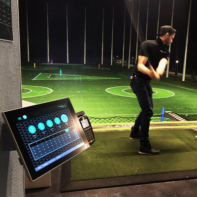 A man mid swing at a golf ball at the driving range, with a bright tablet kiosk in the foreground.
