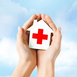 Cupped hands in front of a cloudy background holding a small white house with a red cross on it